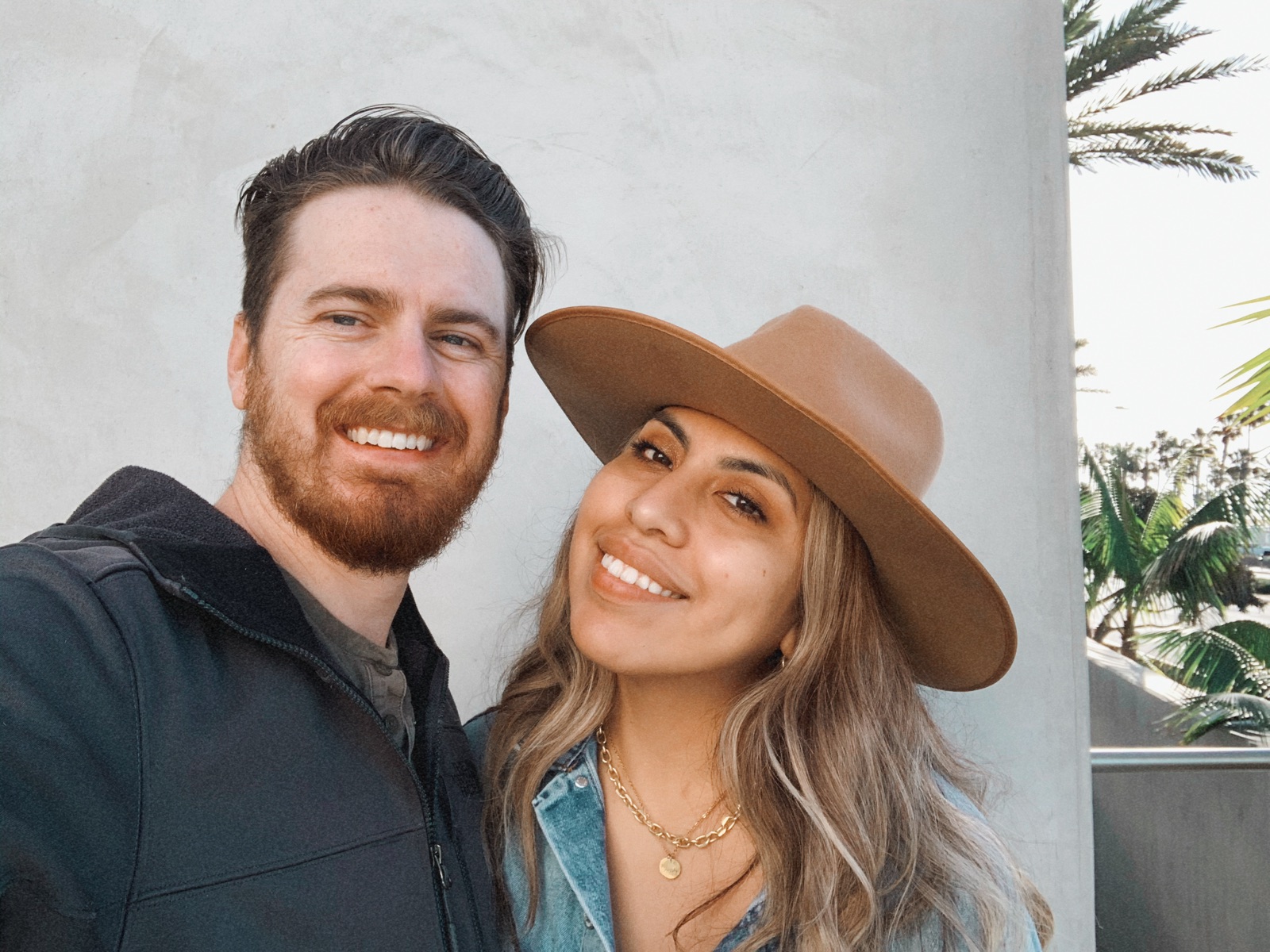 Smiling selfie by a white wall, Adriana in a tan hat